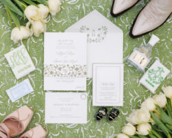 A flat lay detail shot of the invitation suite and personal items of the bride and groom, including bride's shoes and florals